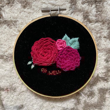 3 pink 3-D embroidered flowers with the words "oh well" rest on black velvet. The embroidery hoops sits on beige carpet
