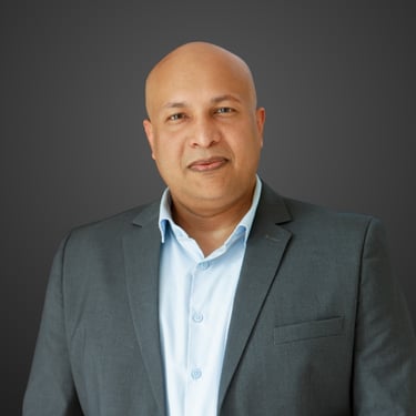 Professional headshot of Simon Gomes, a bald man in a grey suit jacket and light blue shirt.