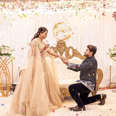 Groom kneeling and holding bride’s hand on wedding stage with confetti falling in celebration