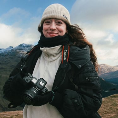 A photographer posing and smiling at the camera, holding her own camera, with mountain ranges in the background.  