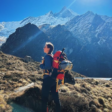 A female hiker standing on a mountain. Behind her is mountain range with ice caps covering the top. 