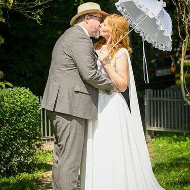 Küssendes Brautpaar im Grünen mit weißem Spitzenschirm nach der Hochzeit.