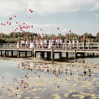 Hochzeitsgesellschaft lässt rote Luftballons auf einem Steg am See steigen – Gruppenmoment einer Hochzeitsreportage.