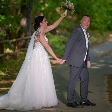 Brautpaar geht Hand in Hand einen Waldweg entlang, die Braut hält den Brautstrauß erhoben.