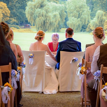 Brautpaar sitzt während der Trauung an einem Tisch, umgeben von Gästen bei einer freien Trauung im Grünen.