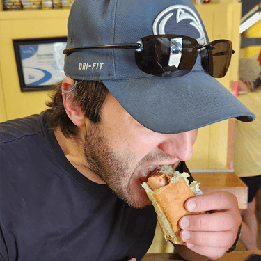 A man in a Penn State baseball cap eating a delicious hot dog indoors.