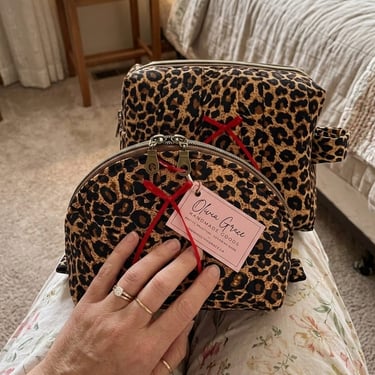 Two leopard print quilted makeup bags with red ribbons and brand tags resting on a person's lap.