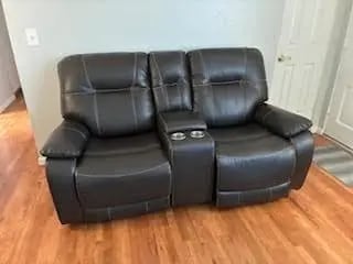 A dark brown leather double recliner loveseat with a center console and cup holders on a light wood floor.