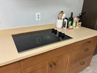 A black glass-top electric stovetop set in a tan countertop with wooden kitchen cabinets and cooking supplies.