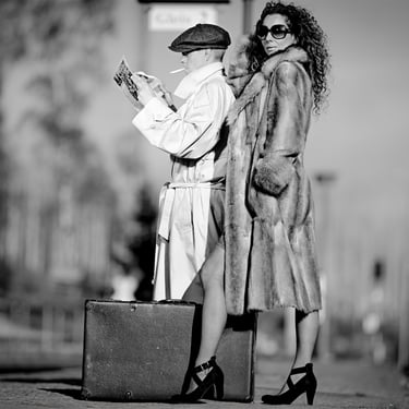 Schwarzweiß-Editorial-Portrait eines Mannes mit Zeitung und einer Frau im Pelzmantel vor urbanem Hintergrund.