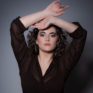 Ein Porträt einer Frau mit dunklen, lockigen Haaren und roten Lippen. Sie trägt eine durchsichtige, dunkelbraune Bluse und ha