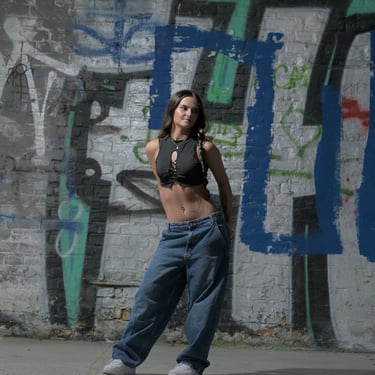 Ganzkörper-Porträt einer jungen Frau in Streetwear vor einer mit Graffiti besprühten Backsteinwand.