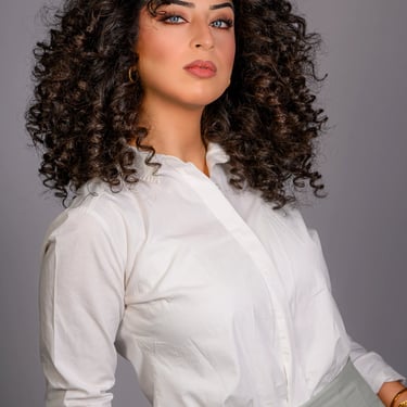  Porträt einer Frau mit Locken, blauen Augen und weißem Hemd, die über die Schulter blickt.