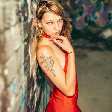 Junge Frau im roten Kleid mit Tattoo am Arm lehnt an einer Graffiti-Wand und blickt direkt in die Kamera – urbanes Portrait v