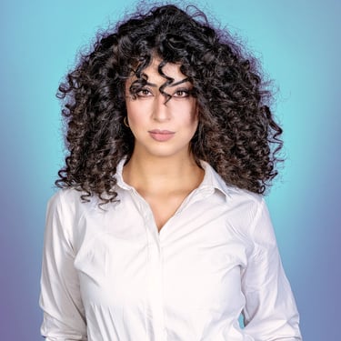 Porträt einer Frau mit voluminösen Locken und einem weißen Hemd vor blauem Hintergrund.