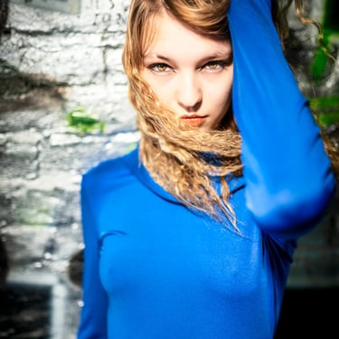 Künstlerisches Portrait einer jungen Frau mit blondem Haar im leuchtend blauen Kleid, intensiver Blick in die Kamera vor urba