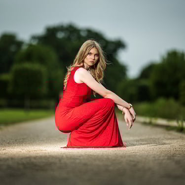  Eine junge Frau im roten Abendkleid hockt auf einem Weg mit Bäumen im Hintergrund.