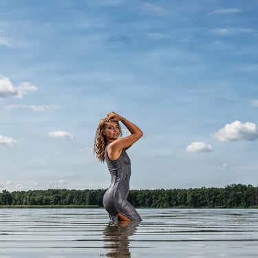 Eine Frau in einem nassen silbernen Kleid steht im See und posiert vor einem bewölkten Himmel.