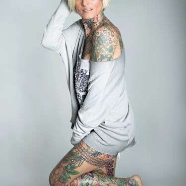 Eine voll tätowierte Frau mit blondem Bob und blaugrünen Augen posiert in einem grauen Sweatshirt.