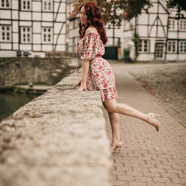 Junge Frau mit roten Haaren und geblümtem Sommerkleid lehnt lächelnd auf einer Steinmauer vor historischen Fachwerkhäusern – 