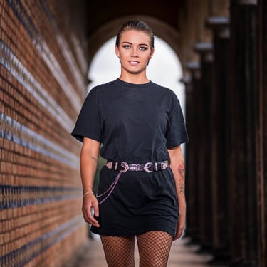 Eine selbstbewusste Frau in schwarzem T-Shirt, Fischernetzstrumpfhose und Gürtel, die in einem gemauerten Gang steht.