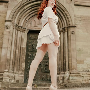 Rothaarige Frau im weißen Kleid posiert selbstbewusst vor einem historischen Kirchenportal mit kunstvollen Steinornamenten – 