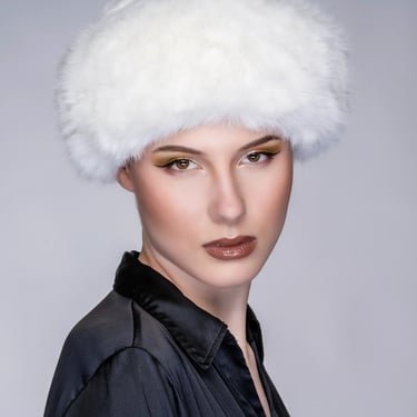  Porträt einer Frau mit weißer Fellmütze, dunkler Bluse und auffälligem Make-up.