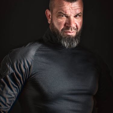 Porträt eines muskulösen Mannes mit Bart und Seitenhaarschnitt in einem schwarzen Rollkragenpullover.