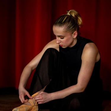 Eine junge Balletttänzerin mit blondem Pferdeschwanz bindet ihre Spitzenschuhe vor einem roten Vorhang.