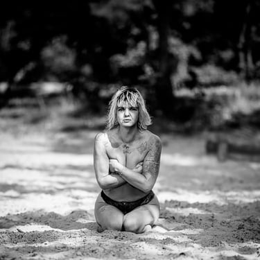 Schwarz-Weiß-Aufnahme einer tätowierten Frau mit kurzen Haaren, die im Sand kniet.