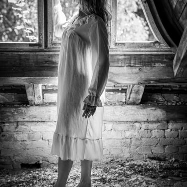 Schwarz-Weiß-Aufnahme einer Frau im Nachthemd, die barfuß an einem Fenster steht.