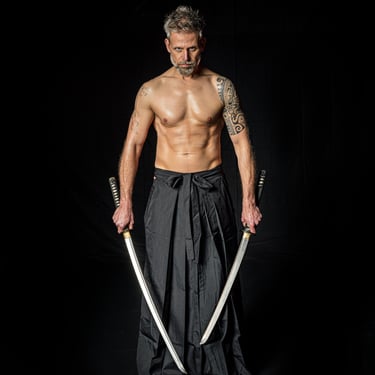 Ein muskulöser Mann mit Tattoos und grauen Haaren, der zwei Schwerter hält und einen schwarzen Hakama trägt.