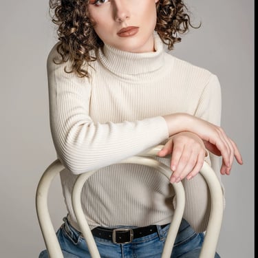 Porträt einer jungen Frau mit lockigem Haar, hellem Rollkragenpullover und Jeans, aufgenommen im Studio von LensAndLook – mod