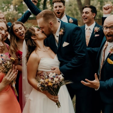 Bride and groom kissing surrounded by cheering wedding party outdoors – captured by Anngye & Juan Photography.