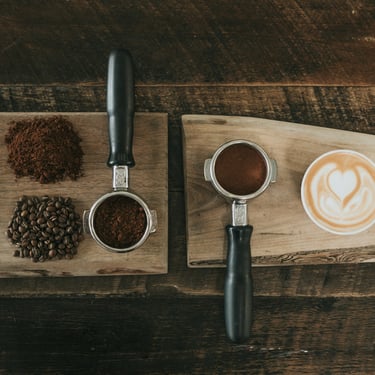 A wooden table with freshly roasted coffee beans, freshly grounded coffee powder, a portafilter and a cup of coffee