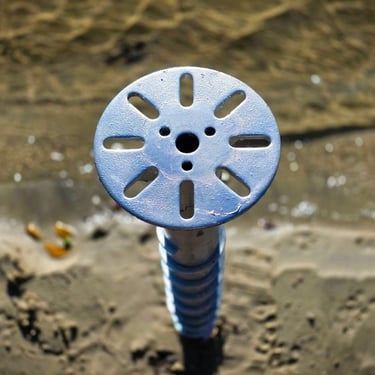 A light blue circular metal screw pile foundation cap installed over beach sand.