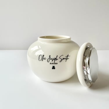 White ceramic urn with soft curves, engraved “Ollie Joseph Smith 25.09.2024” and a teddy bear icon, lid shown open with foil 