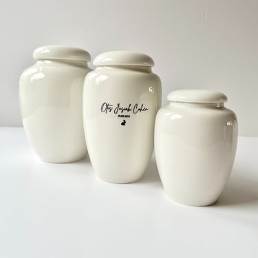 Trio of smooth ivory ceramic urns, centre urn engraved “Otis Josiah Cohen 25.09.2024” with a small black bunny icon.