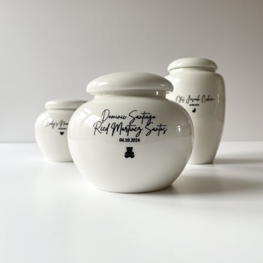 Set of three glossy white ceramic baby urns, each engraved with names, dates, and tiny black heart or bear icons.