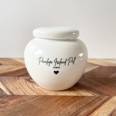 Glossy white ceramic baby urn with “Penelope Ireland Pell 27.09.22” and a heart delicately inscribed in black.