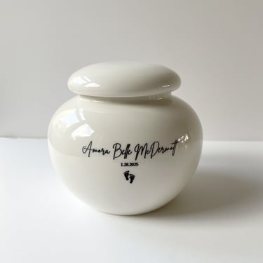 Glossy white ceramic urn with rounded lid, engraved “Amora Belle McDermott, 1.28.2025” above delicate black baby footprints.