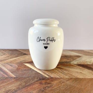 Elegant ivory ceramic baby urn with soft curves, engraved with “Clover Pickles 12.12.2024” above a small black heart.