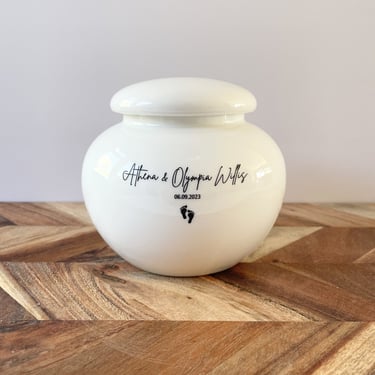 This is a rounded ivory ceramic urn engraved with the text Athena & Olympia Willis, and adorned with a small footprint icon