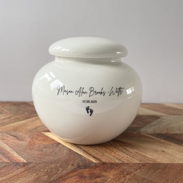 This is a rounded ivory ceramic urn engraved with the text Mason Alan Brooks-Watts, and adorned with a small footprints icon