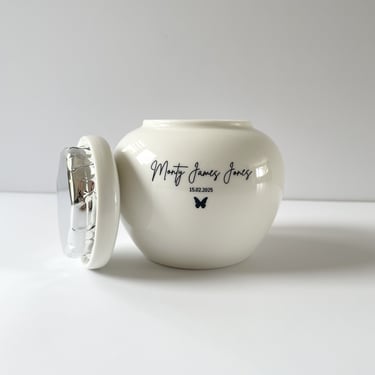 White ceramic urn with soft curves, engraved “Monty James Jones” and a butterfly icon, lid shown open with foil 