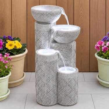 Modern four-tier stone garden water fountain with cascading streams on a patio with potted flowers.
