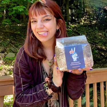 A smiling person holds a Bay Staters mushroom spawn bag outdoors near green foliage.
