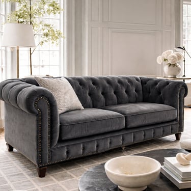 Close up of a charcoal velvet tufted Chesterfield sofa with nailhead trim in a bright classic living room with soft daylight.