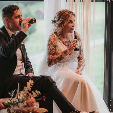 Coole Hochzeiten! Das Hochzeitspaar lauscht mit Drinks in den Händen der Traurede. Der Bräutigam nippt an seiner Bierflasche