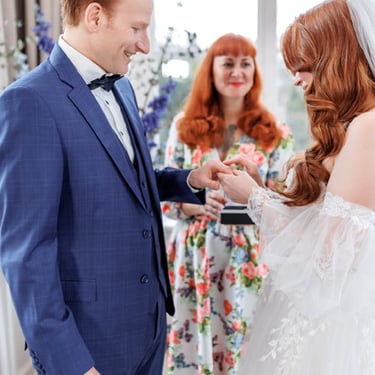 die Braut im romantischen Kleid steckt dem Bräutigam im blauen Anzug den Ring an. Dahinter die lächelnde Traurednerin